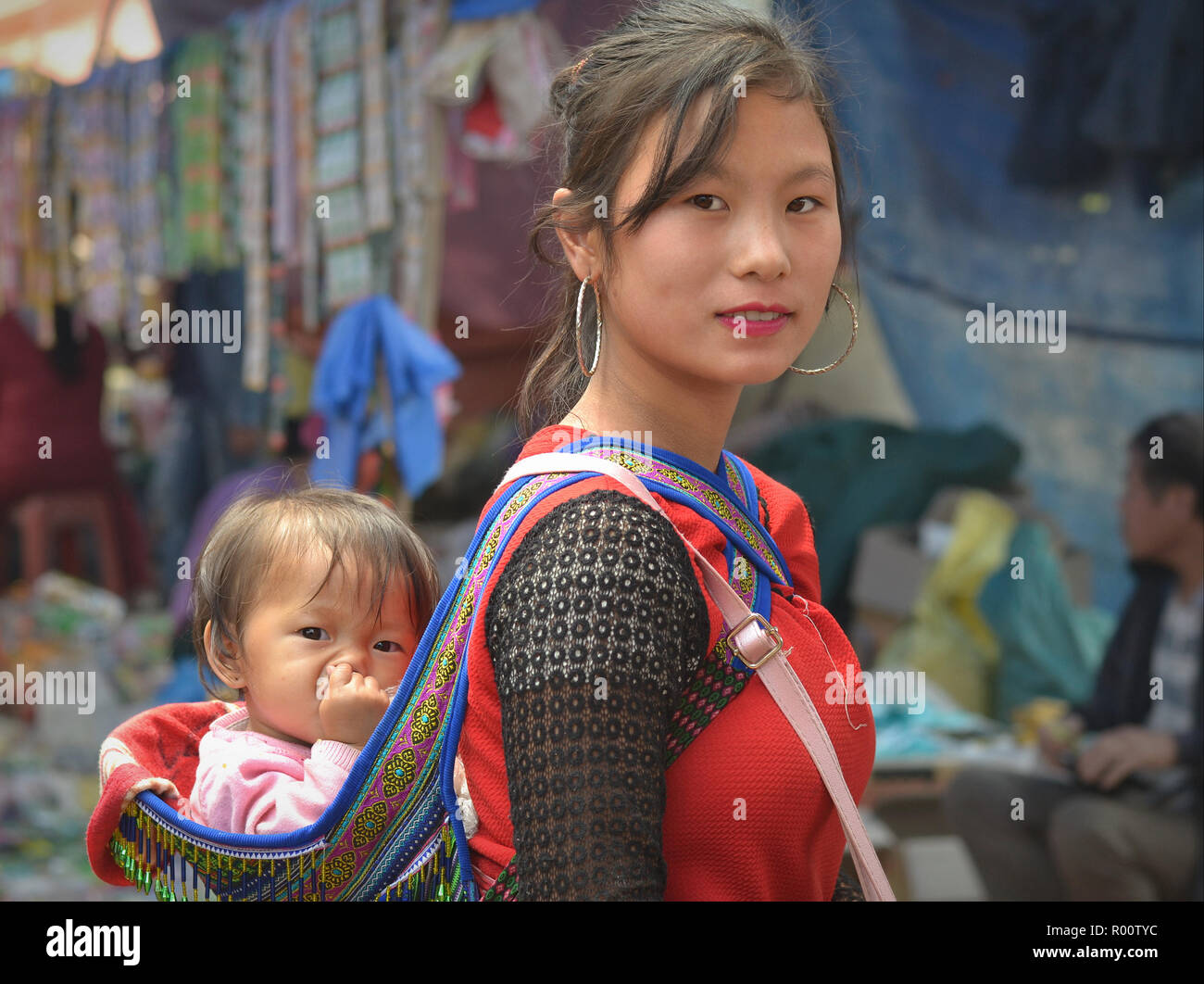 Young Vietnamese Flower H’mong hill-tribe beauty carries on her back a nose-picking baby girl in an embroidered traditional baby sling. Stock Photo