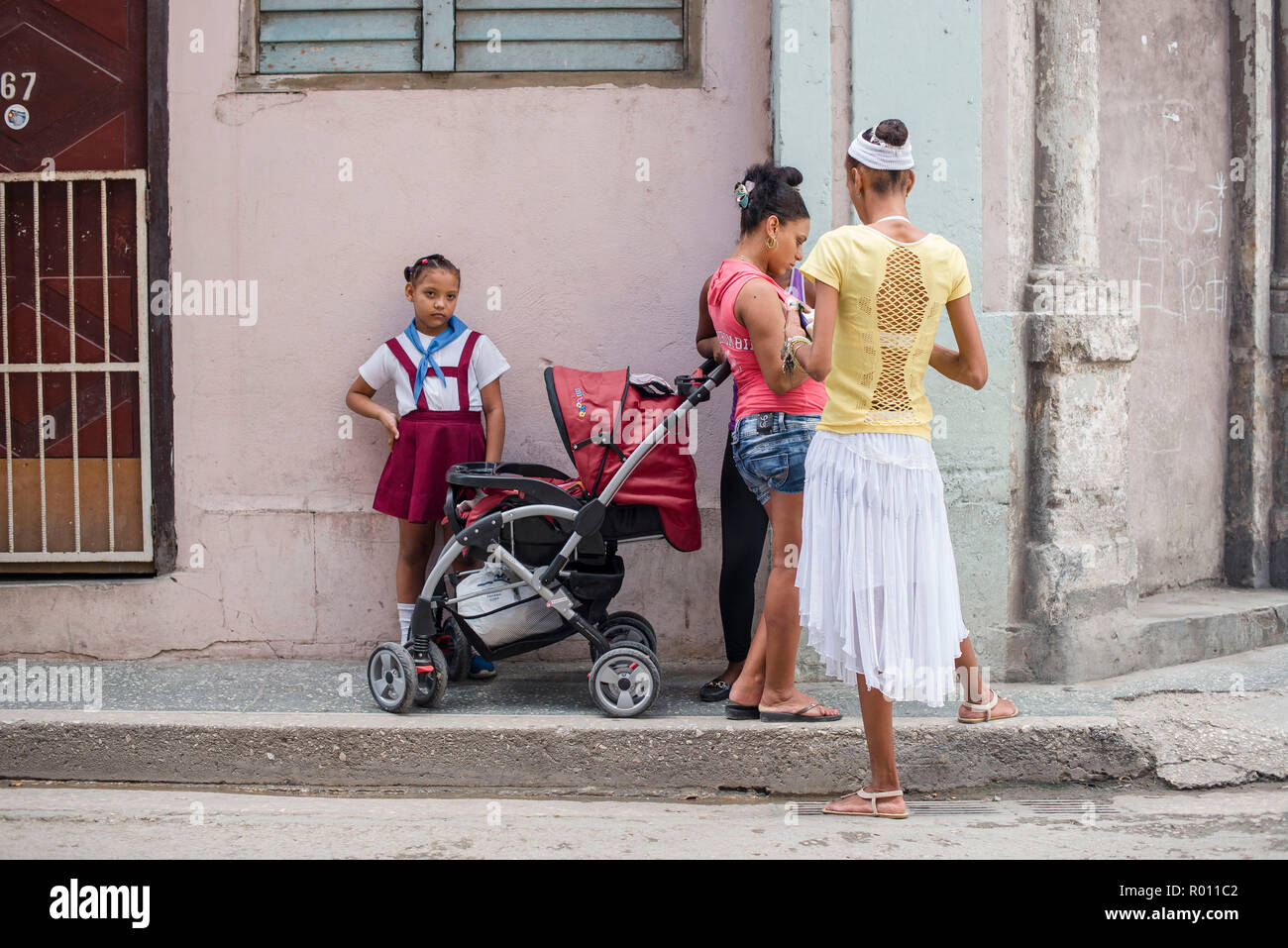 A young girl in a school uniform stands next to a stroller, ignored by the adults around her, on a street corner in Havana, Cuba. Stock Photo