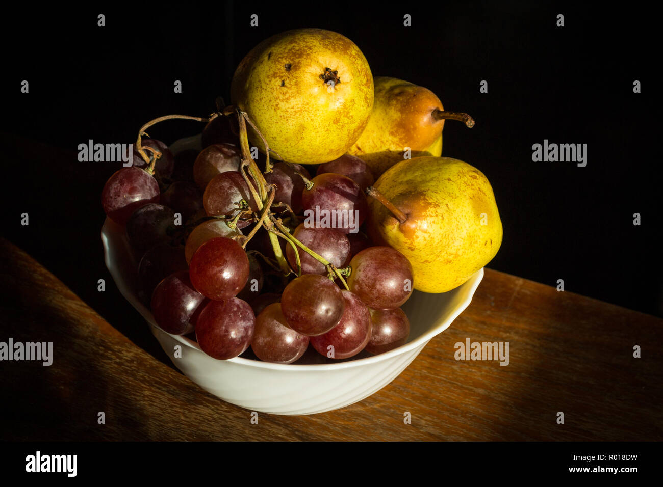 White bowl on brown table containing wine grapes and three pears isolated on black background. Stock Photo