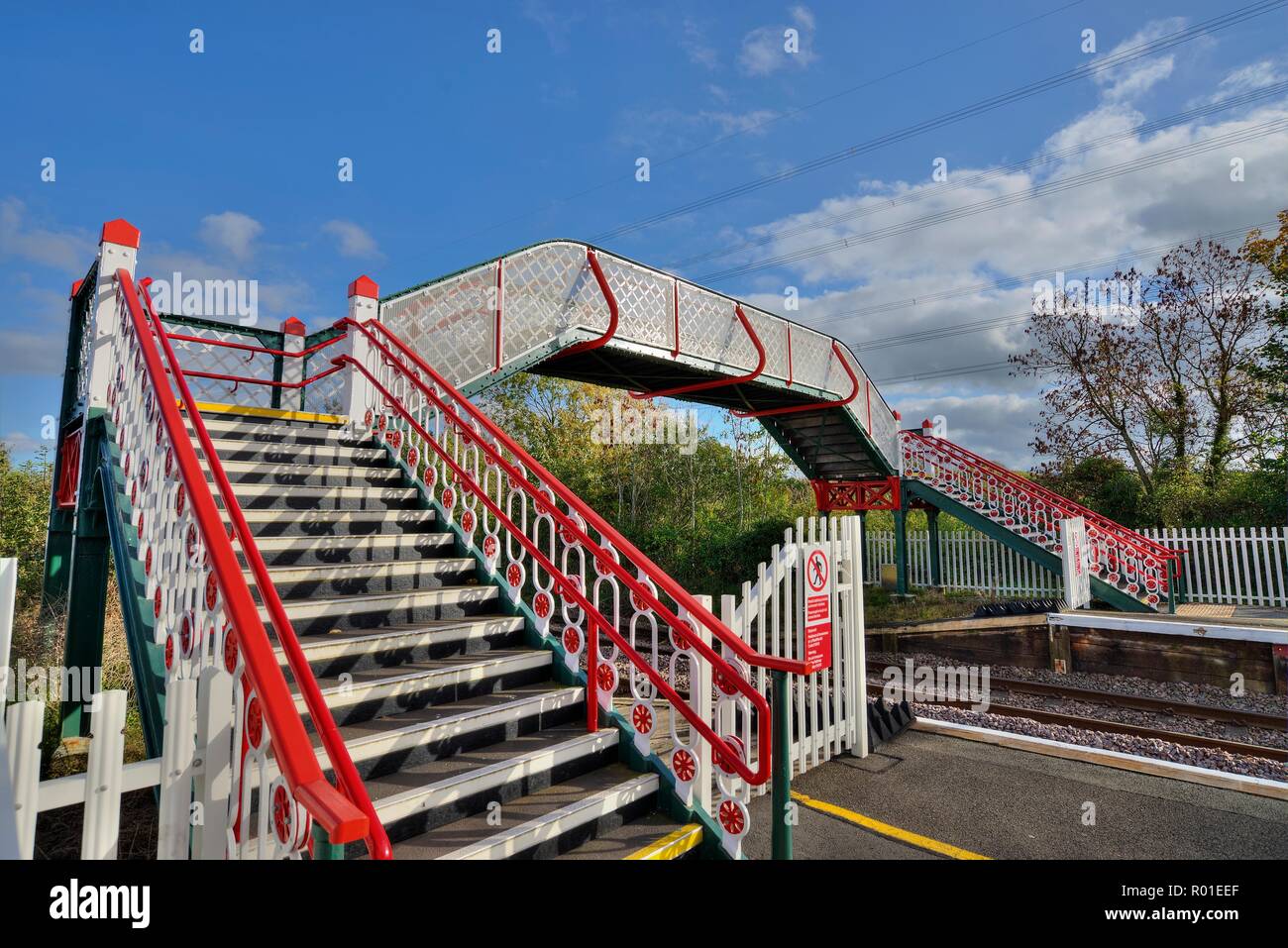 A colourful railway station footbridge at Llanfairpwllgwyngyll, Anglesea in Wales.   Trees are in the background and a blue sky with clouds overhead. Stock Photo