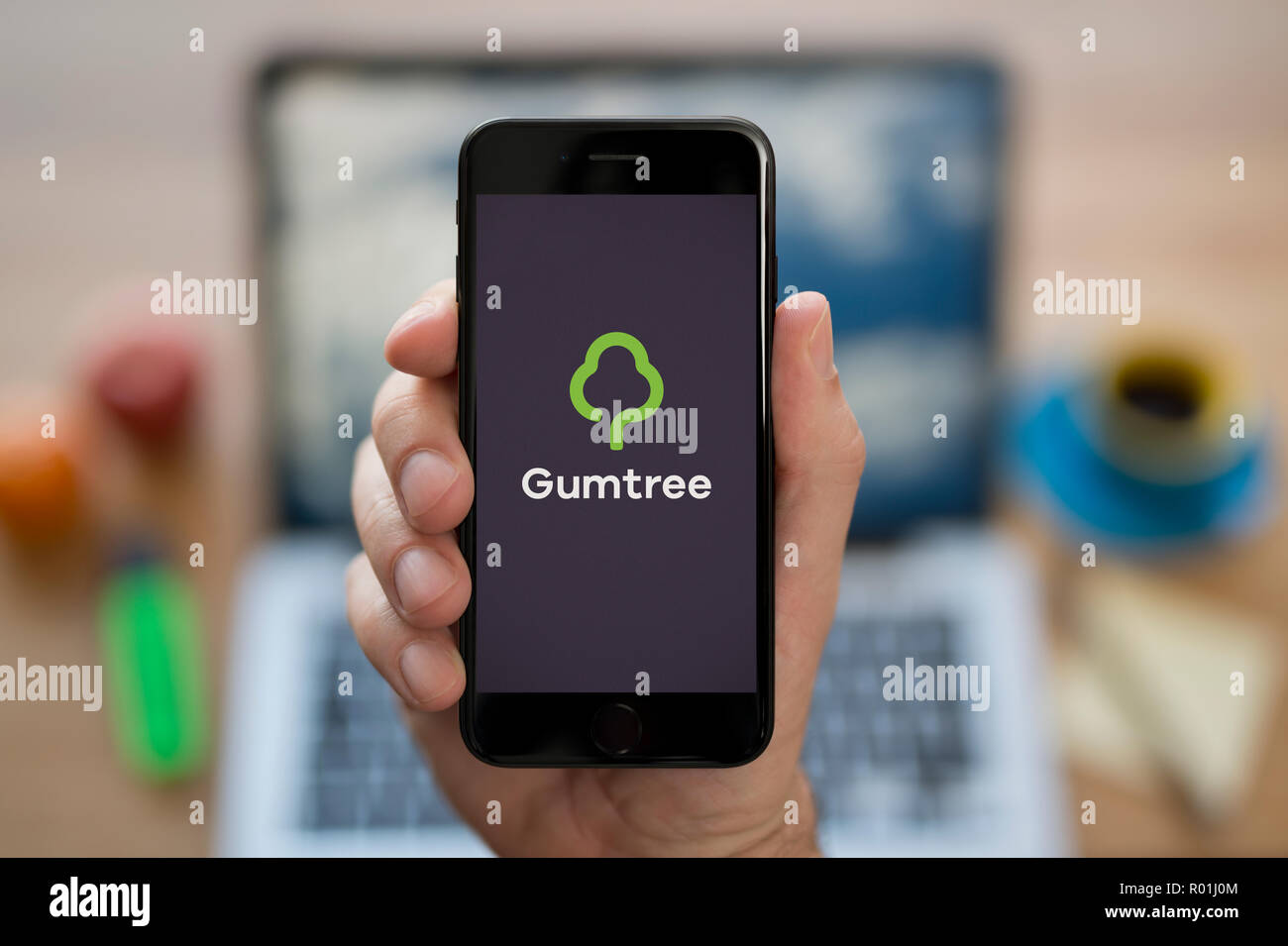 A man looks at his iPhone which displays the Gumtree logo, while sat at his computer desk (Editorial use only). Stock Photo
