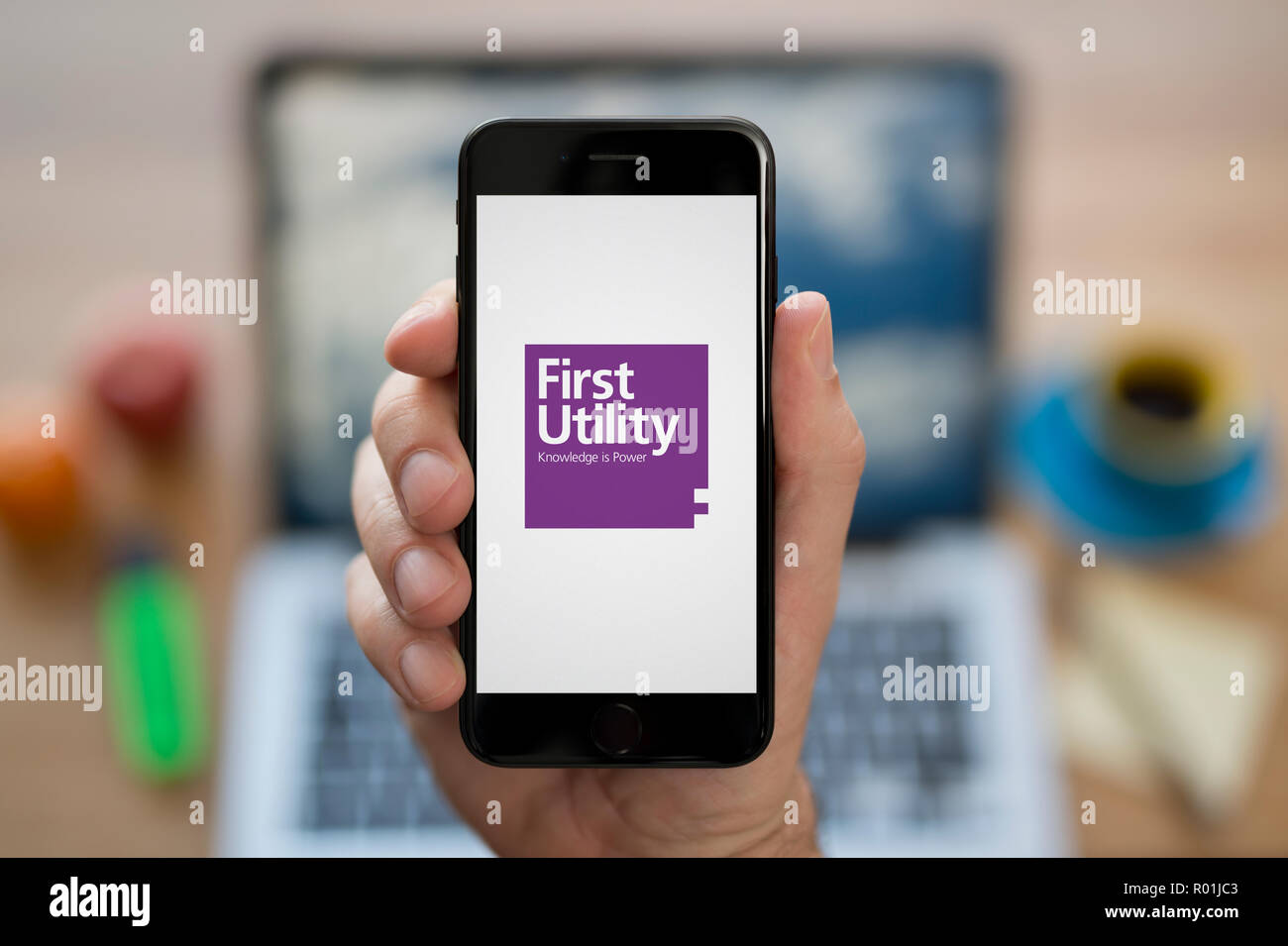 A man looks at his iPhone which displays the First Utility logo, while sat at his computer desk (Editorial use only). Stock Photo