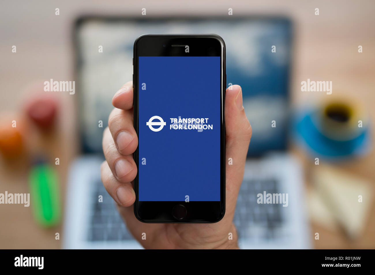 A man looks at his iPhone which displays the Transport for London (TfL) logo, while sat at his computer desk (Editorial use only). Stock Photo