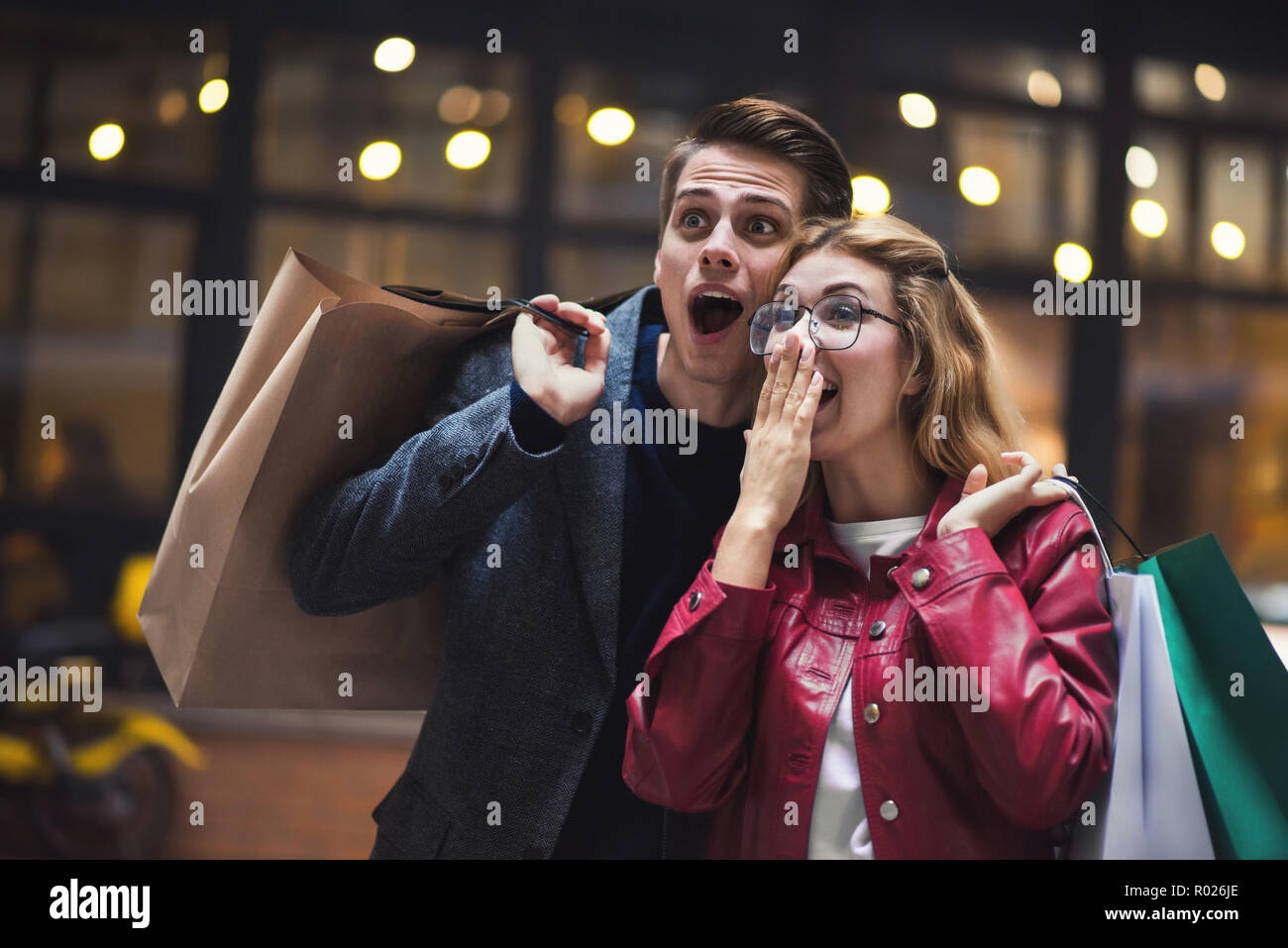Beautiful couple with shopping bags is talking and smiling while doing shopping in the mall focus on the surprised woman. Stock Photo