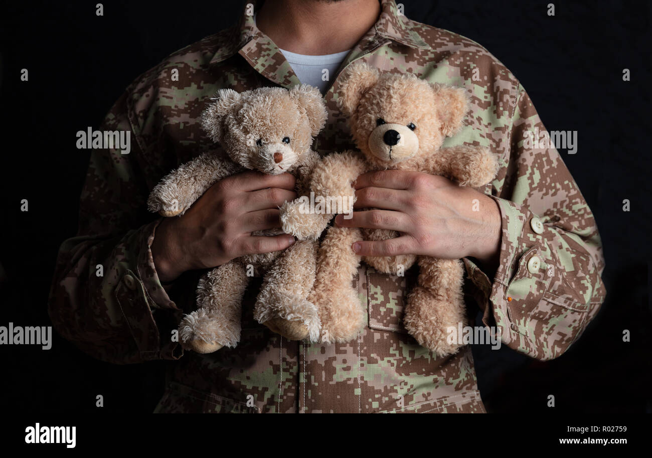 Father in the Army. Young soldier holding two teddy bears standing on black background Stock Photo