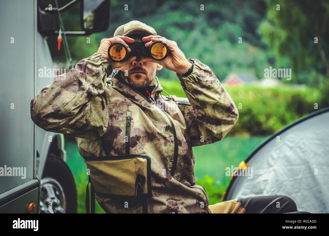 Hunting Season Game Spotting. Caucasian Hunter with Binoculars and the Campsite in Background. Stock Photo