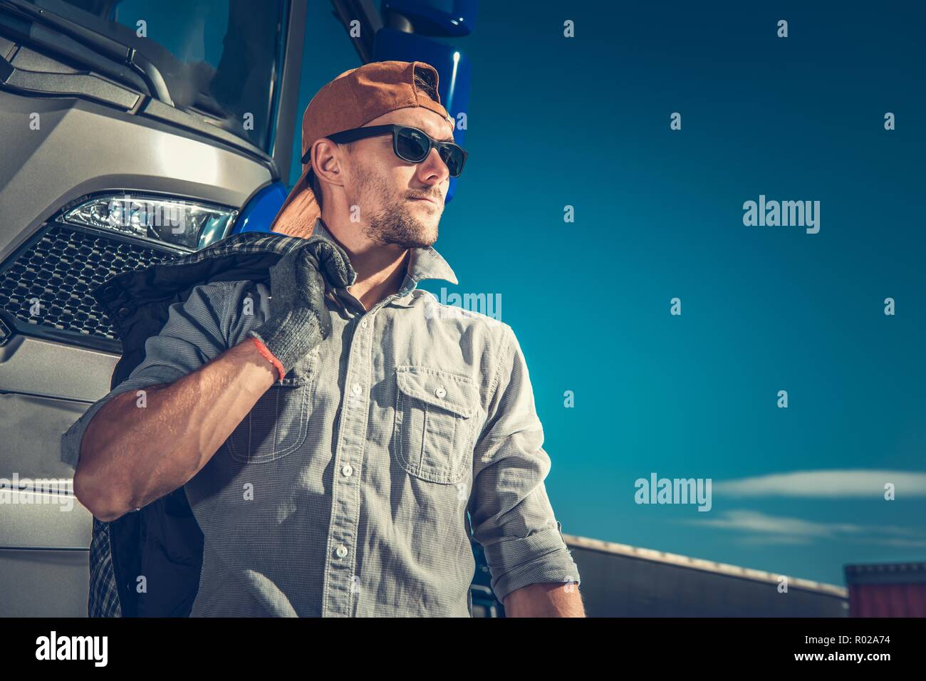 Relaxed Caucasian Truck Driver Wearing Baseball Hat and Sunglasses. Semi Truck in the Background. Stock Photo