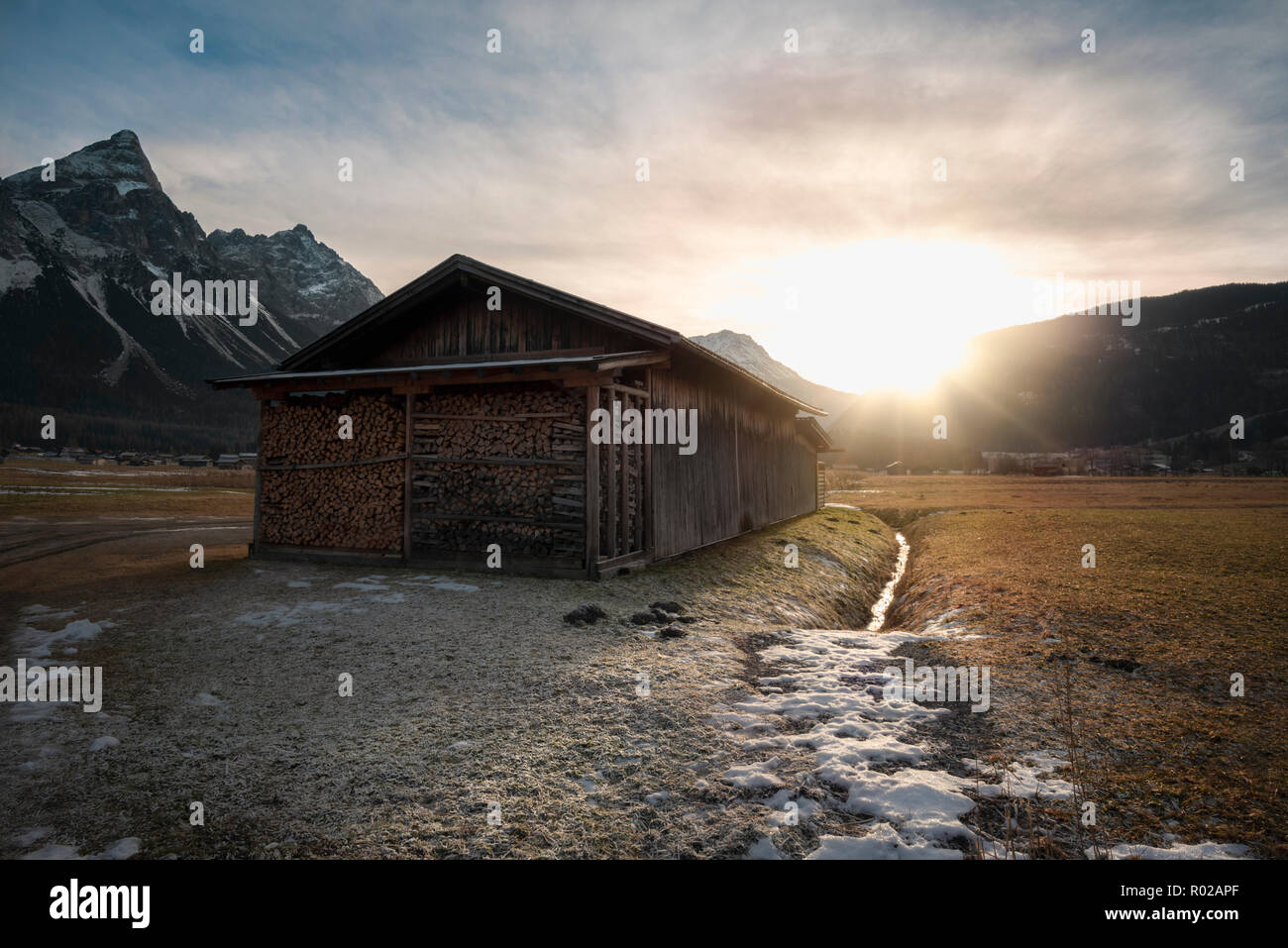 Wooden Barn With A Pile Of Wood Logs On A Meadow With Snow And