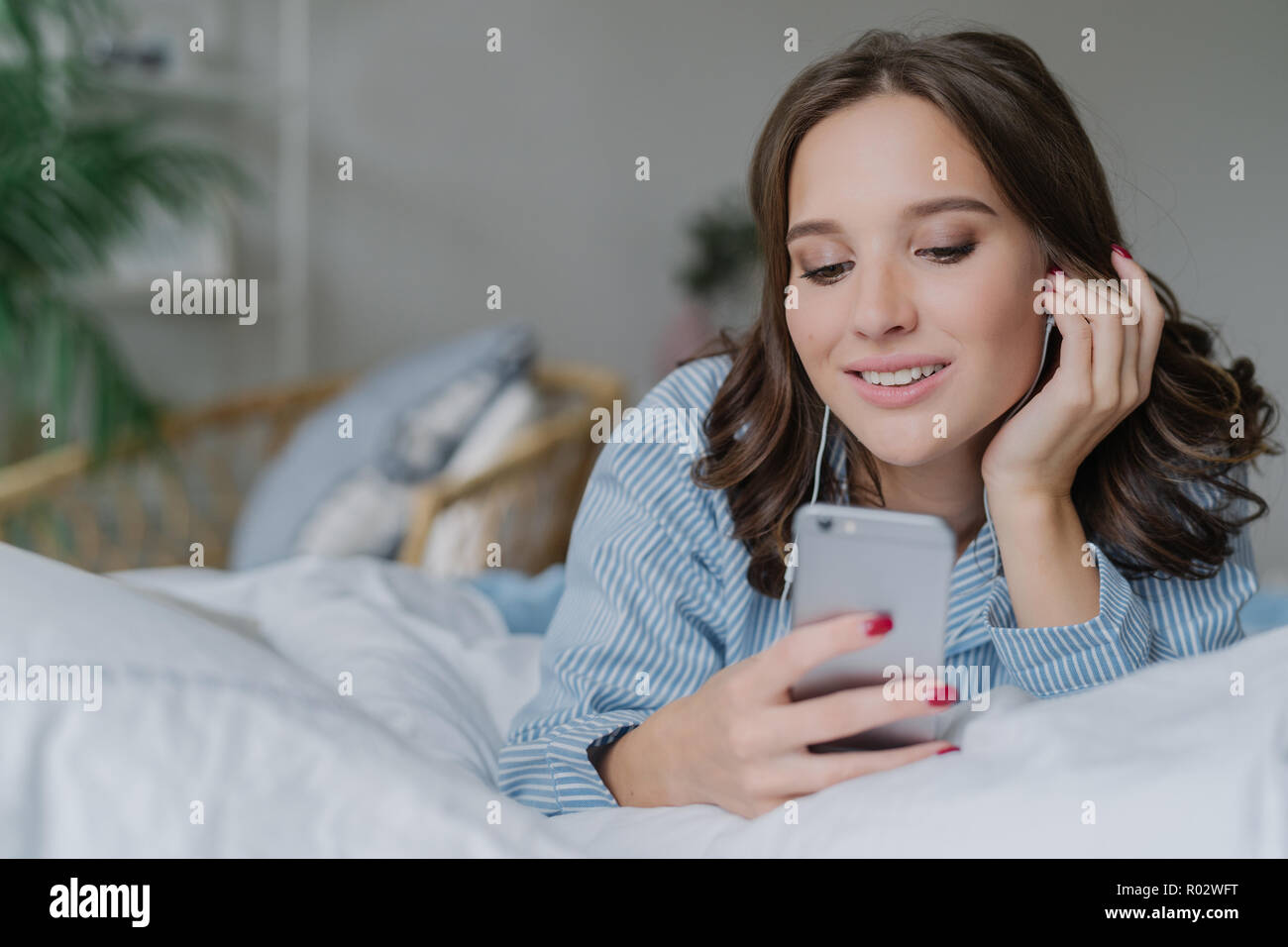 Beautiful woman listens music, makes video call, lies in comfortable bed in morning, dressed casually, has charming smile, poses in bedroom. People, r Stock Photo