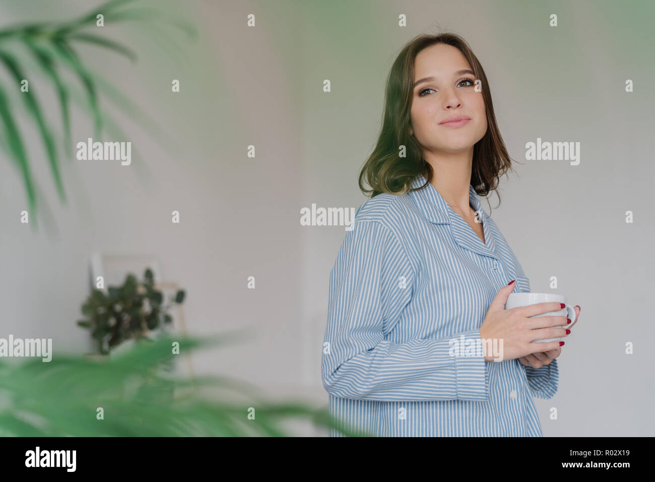 Sideways shot of dreamy Caucasian woman dressed in casual clothes, drinks tea, stands indoor against domestic interior with copy space for your advert Stock Photo