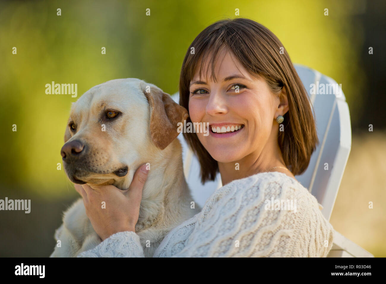 Portrait of a smiling young woman relaxing with her dog on a deck chair in a sunlit garden. Stock Photo
