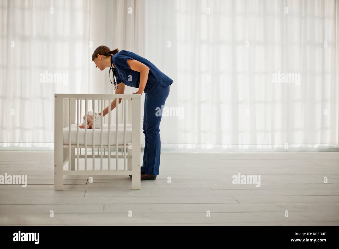 Young nurse bending over a sleeping baby's cot to gently check on him. Stock Photo