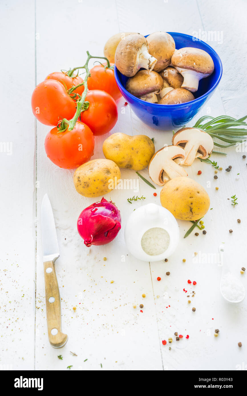 white tabletop displaying brown portobello mushrooms in a blue bowl, tomatoes, potatoes, a red onion, herbs and coarse sea salt and red, green Stock Photo