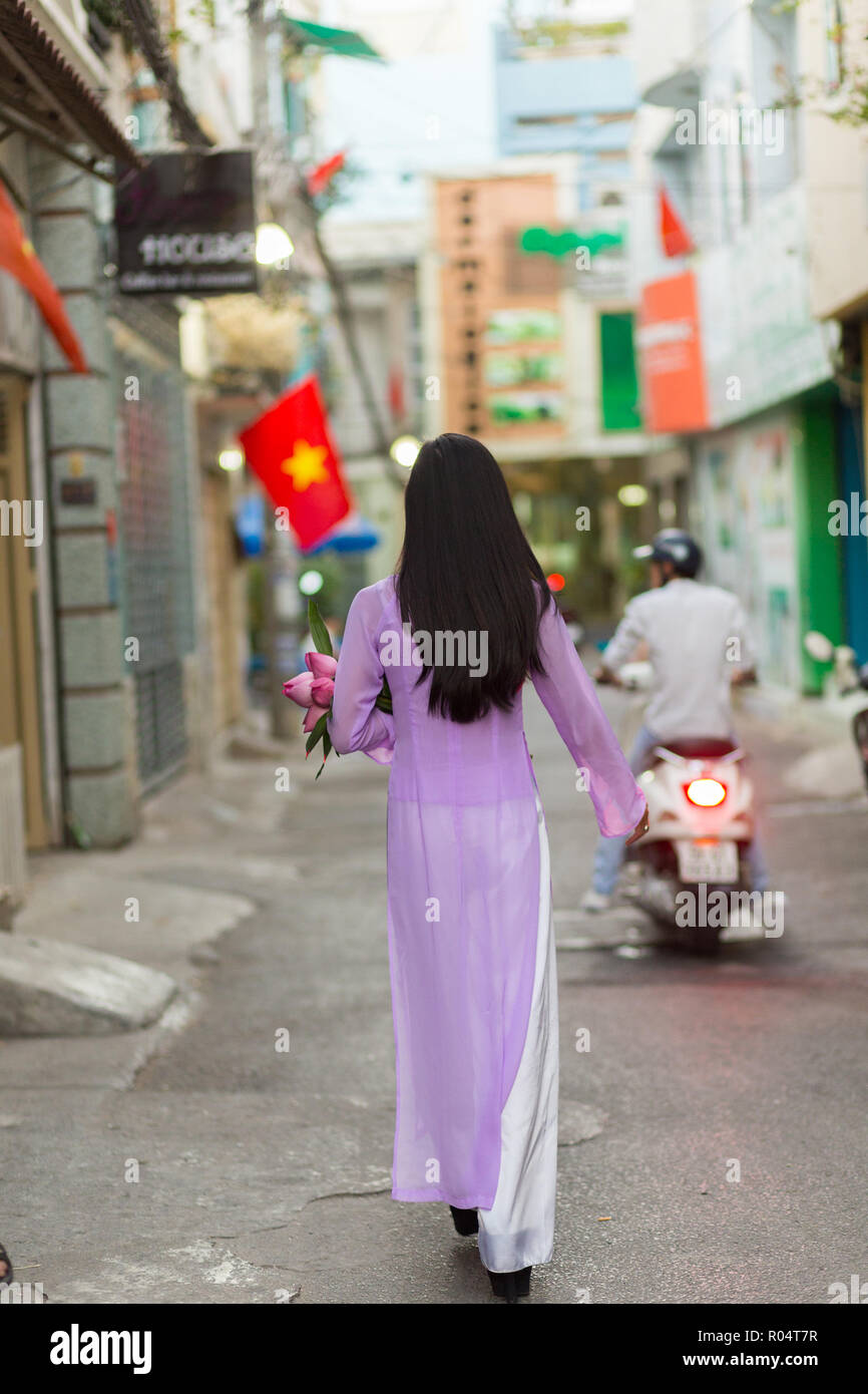 Vietnamese woman walking in Ho Chi Minh street wearing Ao Dai traditional dress, carrying lotus flowers with the Vietnam flag in background . Stock Photo