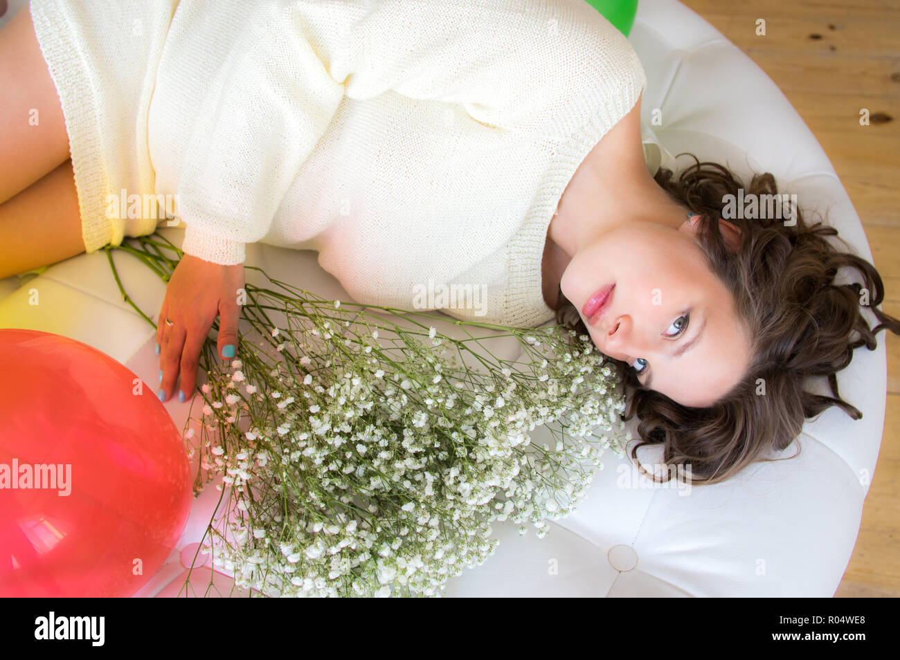 Pregnant woman in white dress lies on round sofa with bouquet of flowers Stock Photo