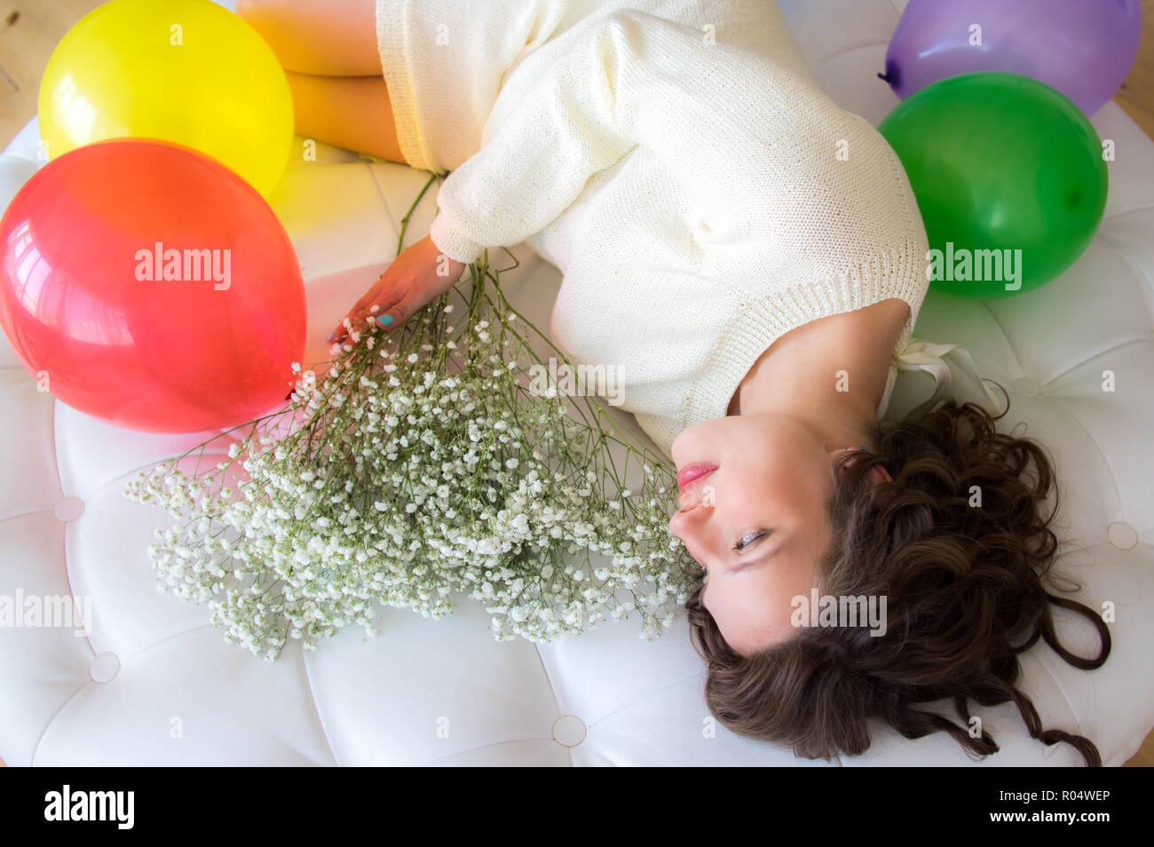 Pregnant woman in white dress lies on round sofa with bouquet of flowers Stock Photo