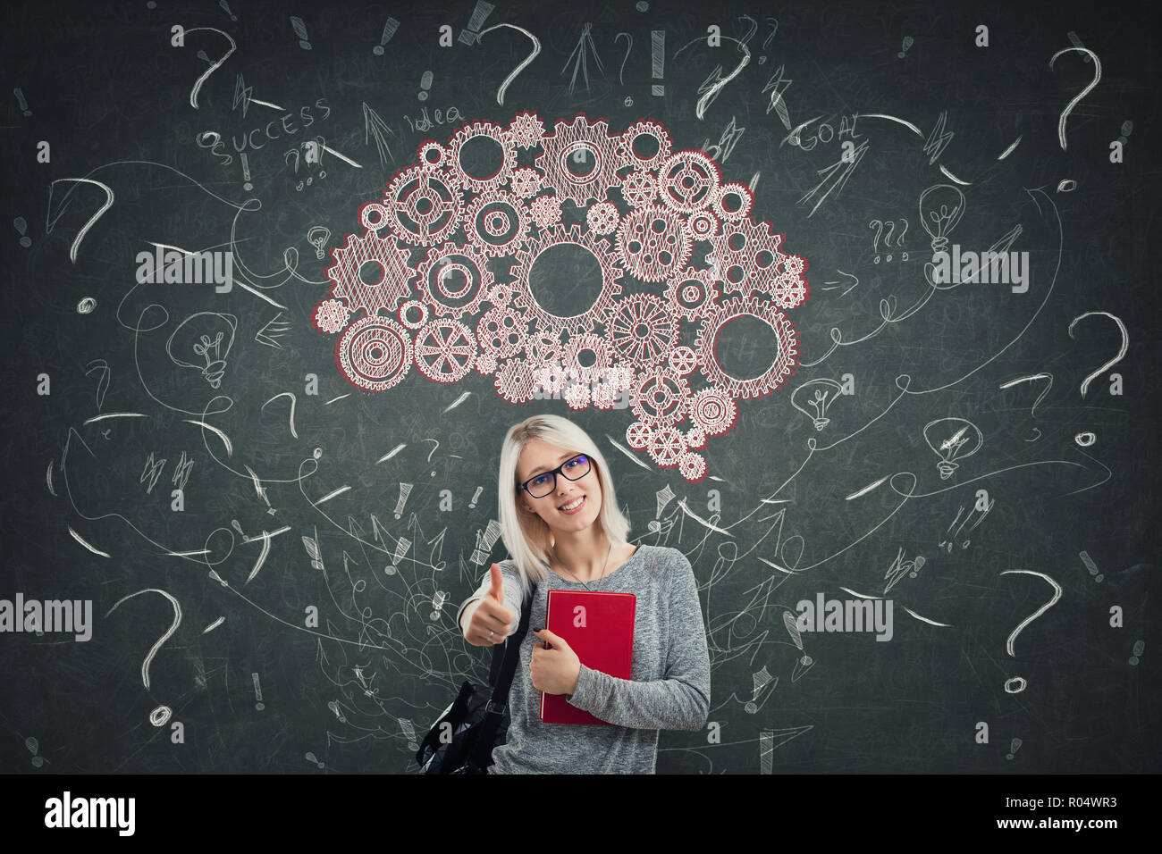 Young woman student wearing glasses holding a book thumb up in front of a blackboard. Gear wheels brain above head, positive thinking mess as thoughts Stock Photo