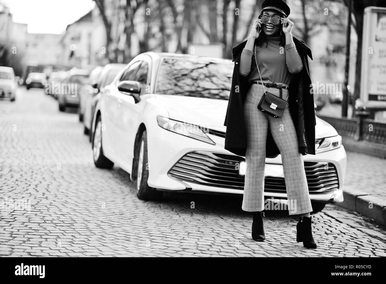 African american fashion girl in coat, newsboy cap and sunglasses posed at street against white business car. Stock Photo