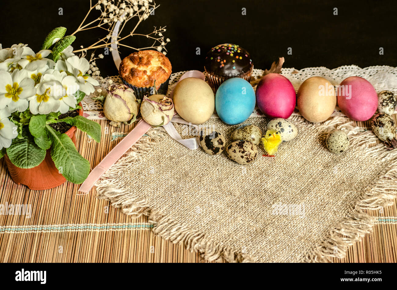 Painted Easter eggs and quail eggs,pastries, a pot of white violet, toy yellow chick with burlap on a straw carpet Stock Photo