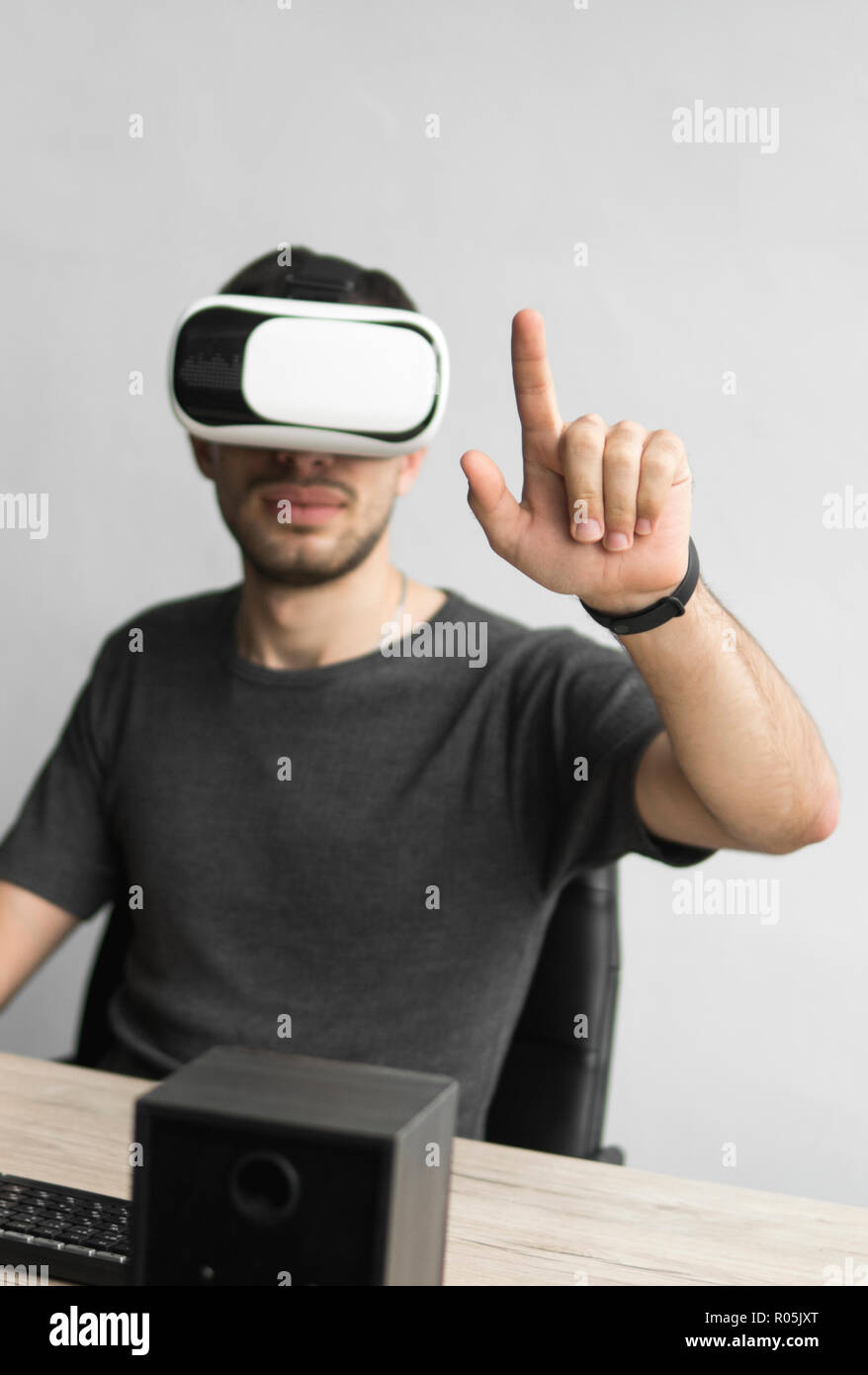 Young man wearing virtual reality goggles headset and sitting in the office against computer. Connection, technology, new generation. Man trying to touch objects or control VR with a hand. Stock Photo