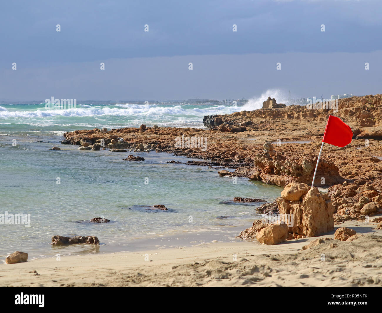 red warning flag on a stony beach during a storm with breaking waves Stock Photo