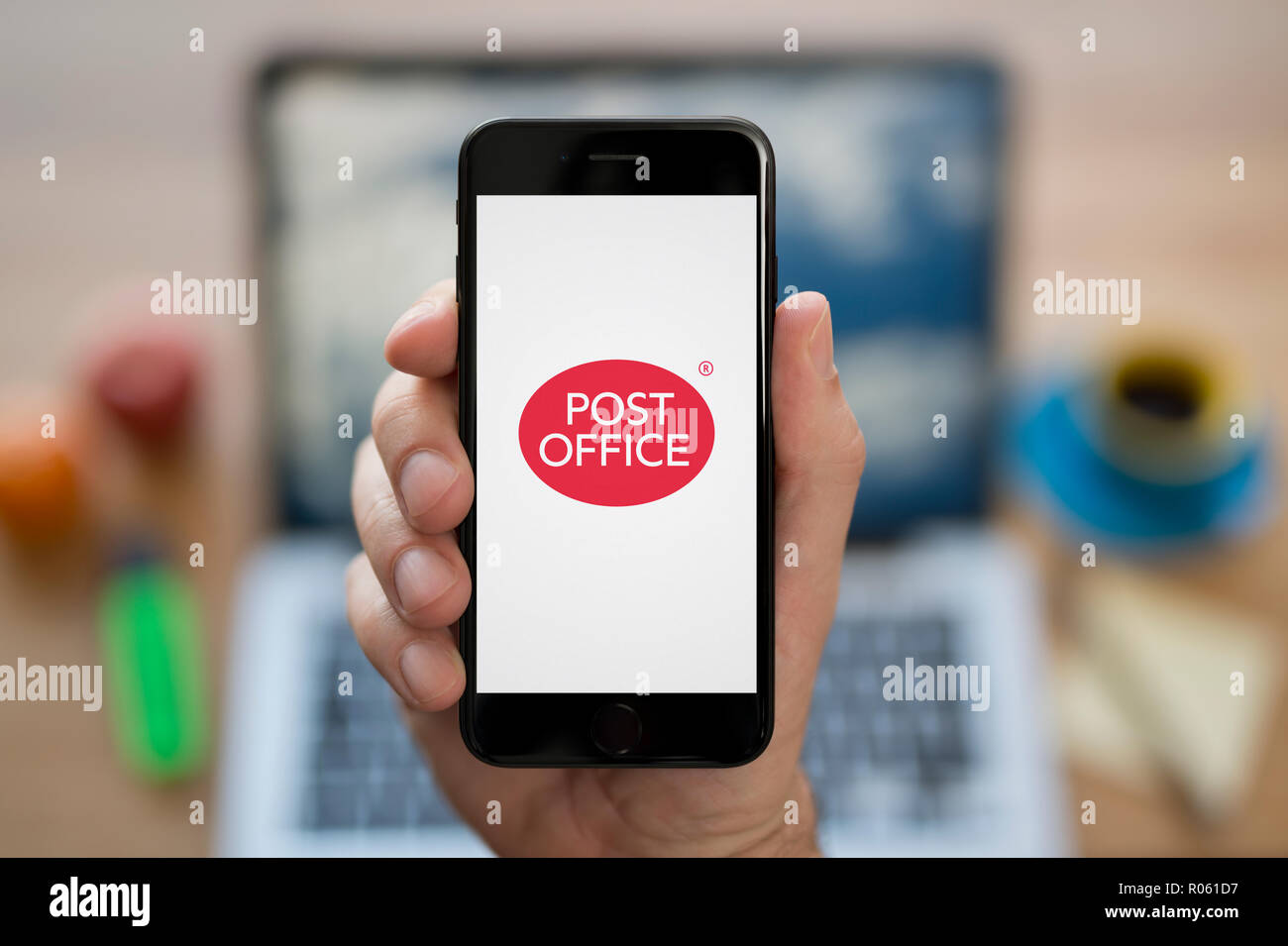 A man looks at his iPhone which displays the Post Office logo, while sat at his computer desk (Editorial use only). Stock Photo