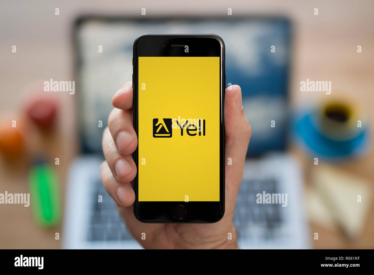 A man looks at his iPhone which displays the Yell logo, while sat at his computer desk (Editorial use only). Stock Photo