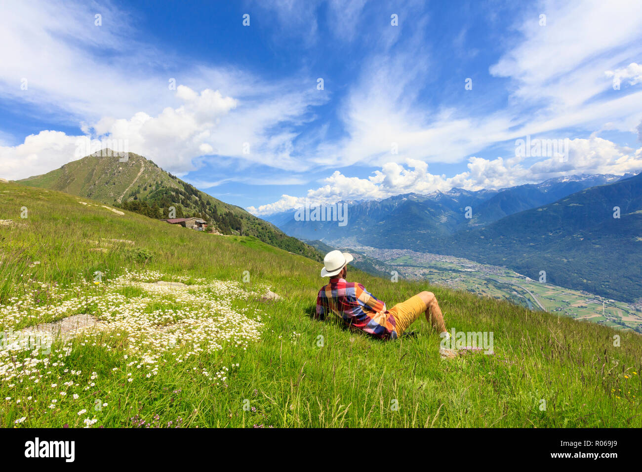 Man with hat relaxes on green meadows looking towards Morbegno, Alpe Bassetta, Valtellina, Sondrio, Lombardy, Italy, Europe Stock Photo