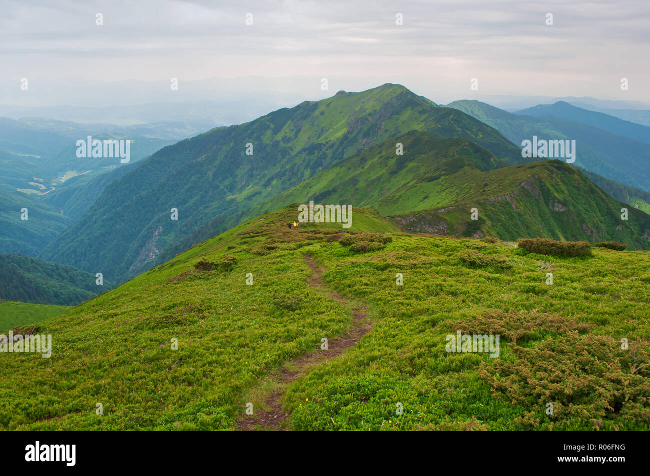 Small figures of travelers descending down a winding path leading to majestic mountain peak and hills covered in green lush grass. Sunny cloudy day in Stock Photo