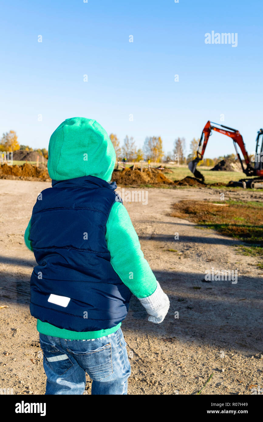 Back view on cute little toddler boy standing on playground and looking at big construction tractor, digging land in rural. Stock Photo