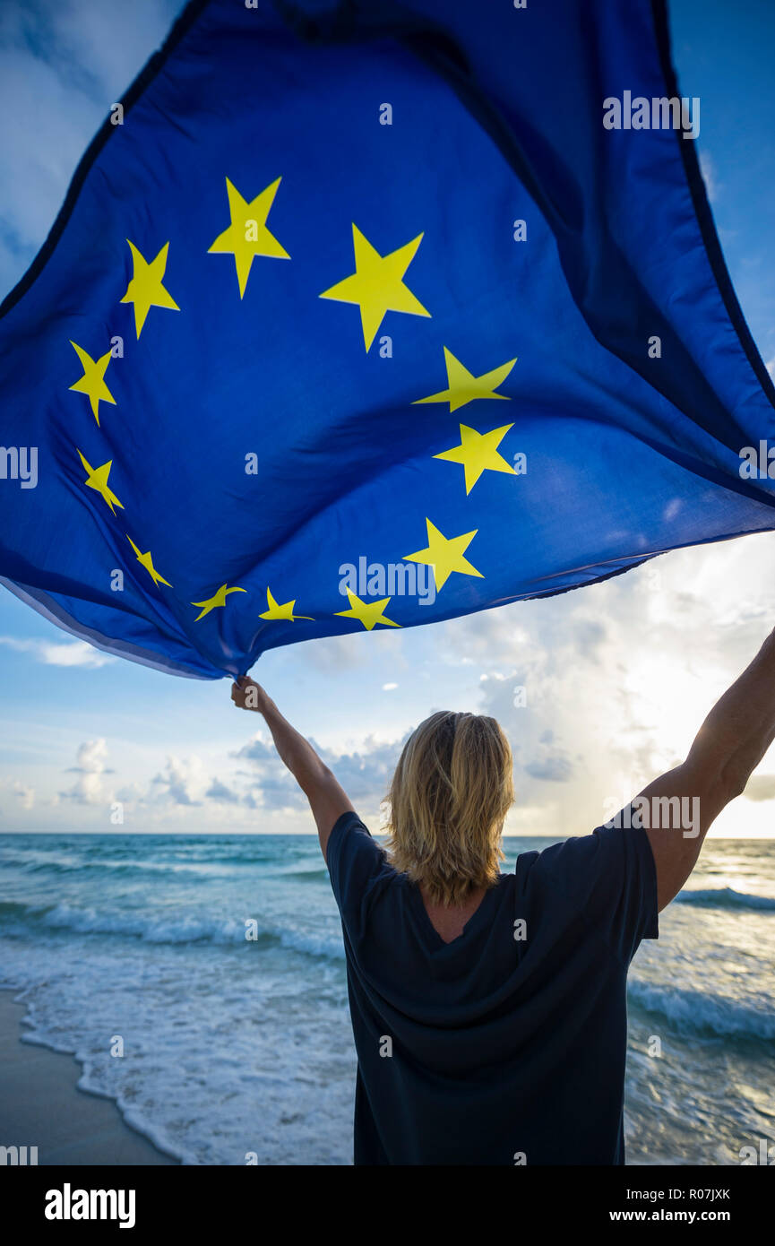 Man with blond hair standing on the shore of a Mediterranean beach holding a European Union EU flag blowing in the wind Stock Photo