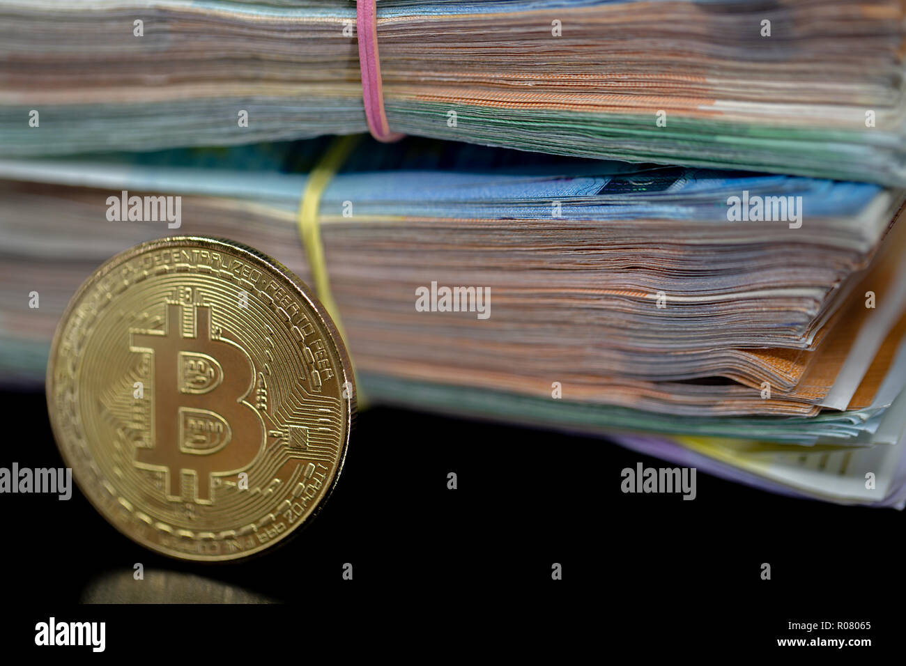 Bitcoin token on black with reflection, with a big pile of euro banknote money in the background Stock Photo