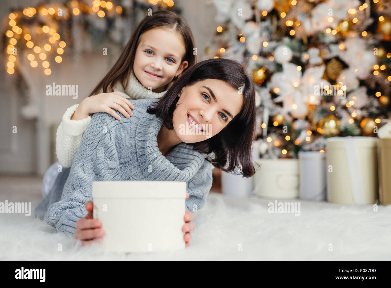 Children, family and celebration concept. Adorable female in knitted sweater holds white present box and small kid stands behind her back, have good m Stock Photo