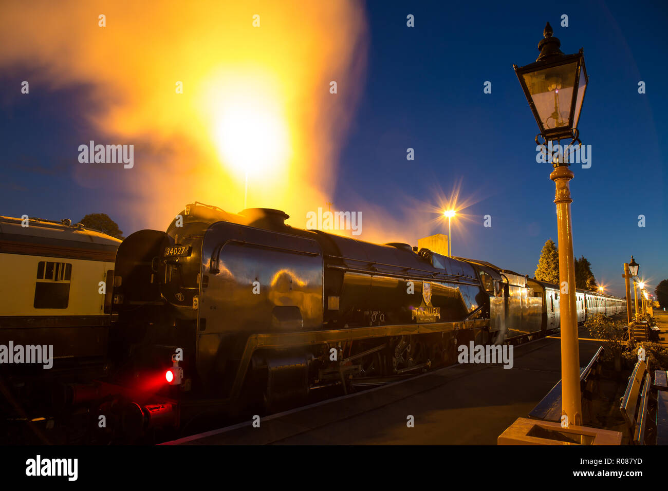 Long exposure, night shot of UK steam locomotive, back-lit, alongside platform, in the dark. Dramatic effect as engine releases steam under pressure. Stock Photo