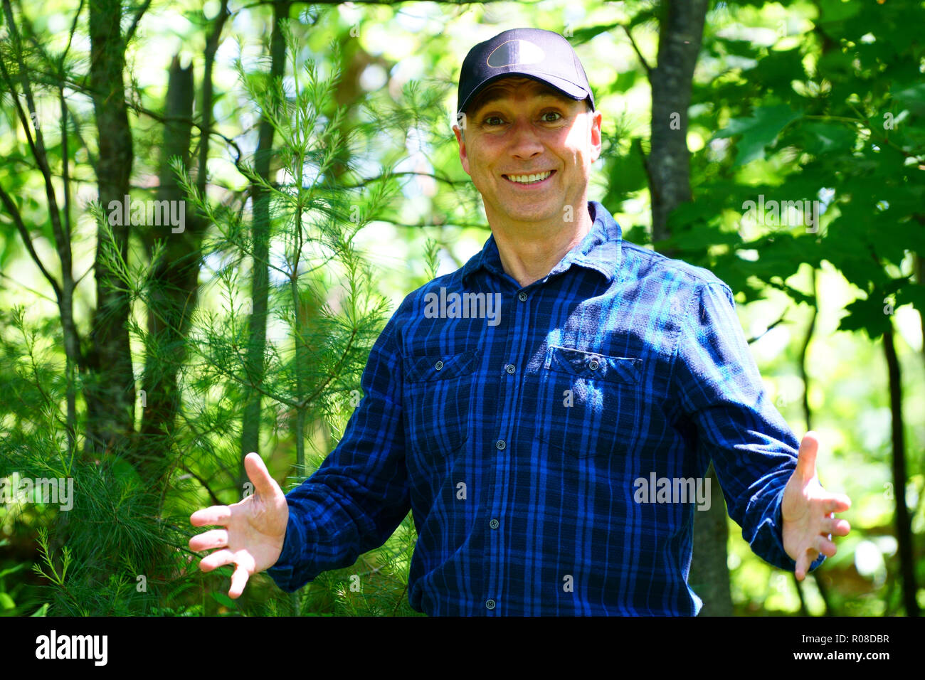A caucasian smiling man is looking into the camera for a portrait while hiking in the forest wearing a blue check shirt and a black hat. Stock Photo