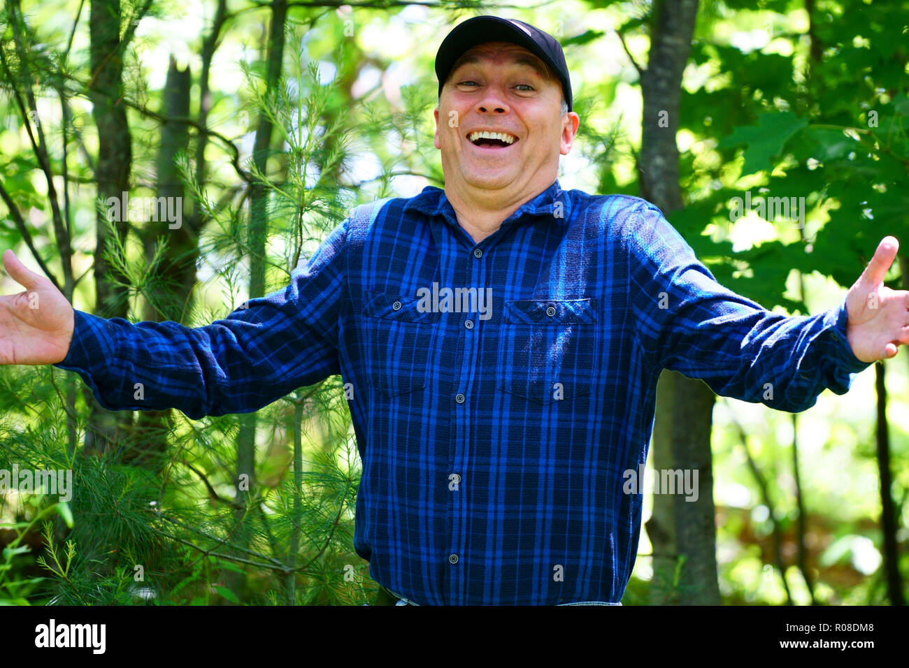 A caucasian smiling man is looking into the camera for a portrait while hiking in the forest wearing a blue check shirt and a black hat. Stock Photo