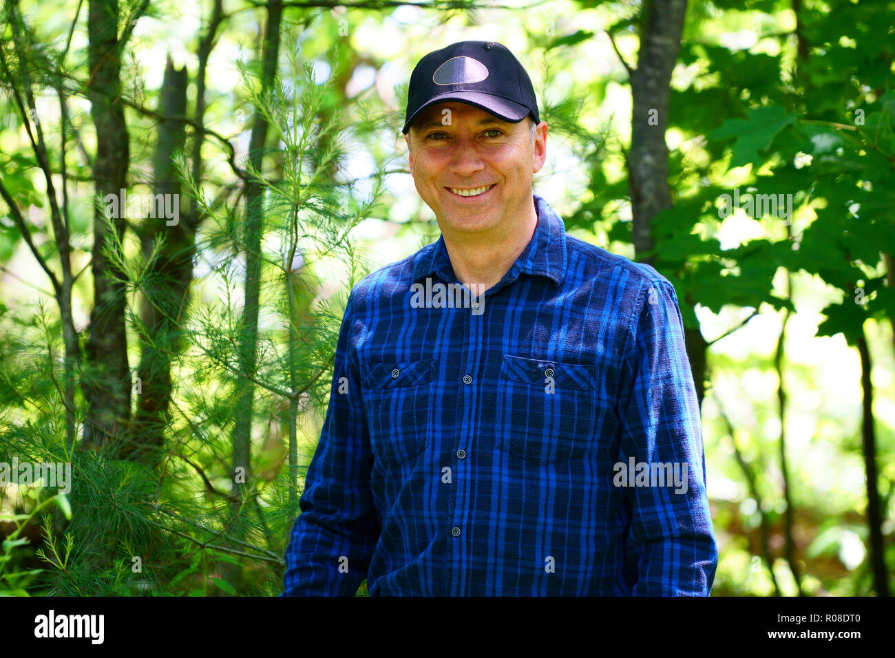 A caucasian smiling man is looking into the camera for a portrait while hiking in the forest wearing a blue check shirt and a black hat. Stock Photo