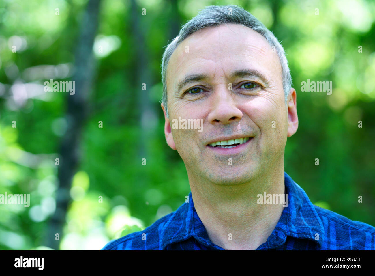 A portrait of a caucasian smiling man looking into the camera while hiking in the forest wearing a blue check shirt with no hat. Stock Photo