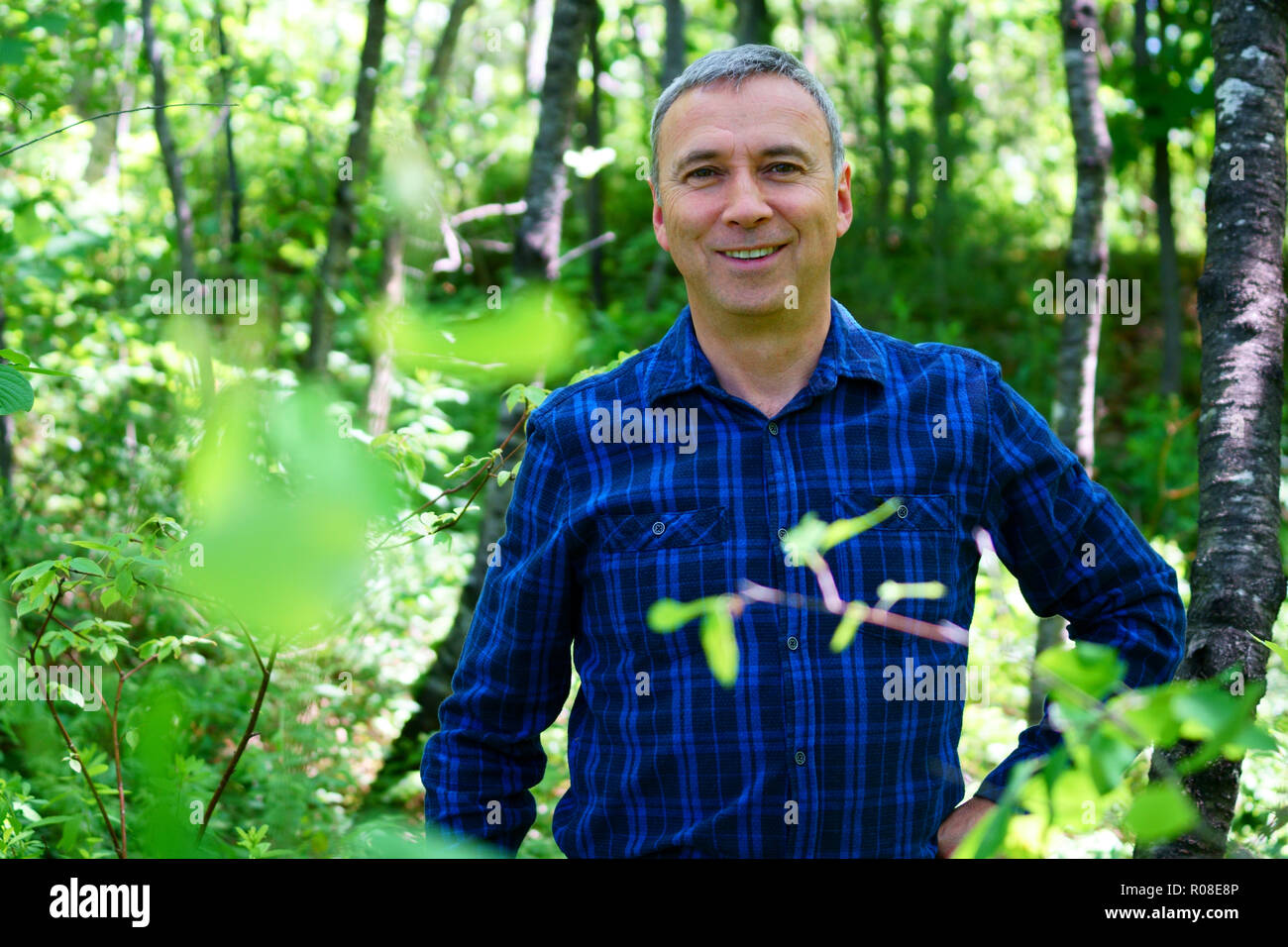A caucasian smiling man is looking into the camera for a portrait while hiking in the forest wearing a blue check shirt and no hat. Stock Photo