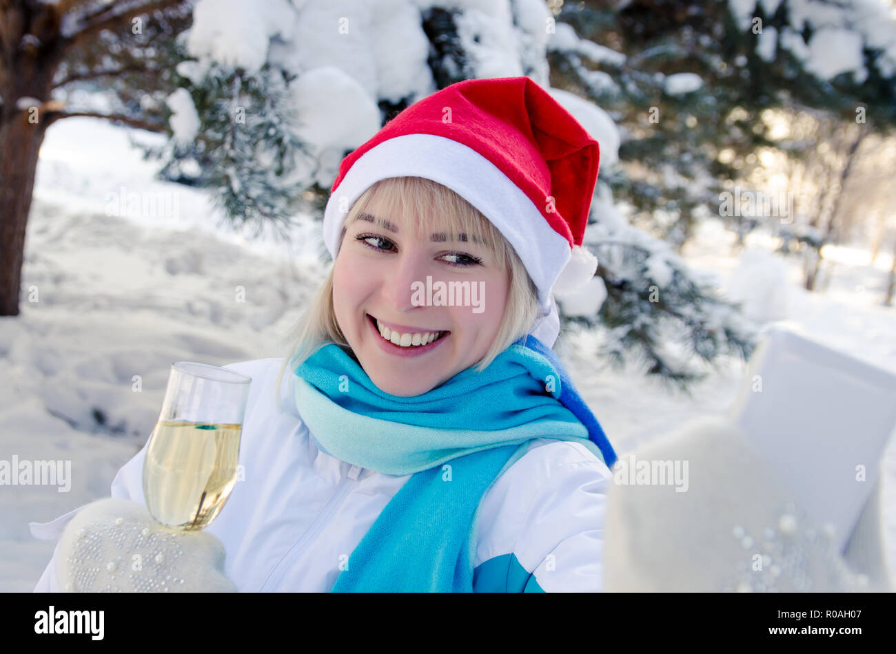 Beautiful attractive blonde in a red Christmas hat with a glass of champagne in her hands celebrates the New Year and takes a selfie on a smartphone Stock Photo
