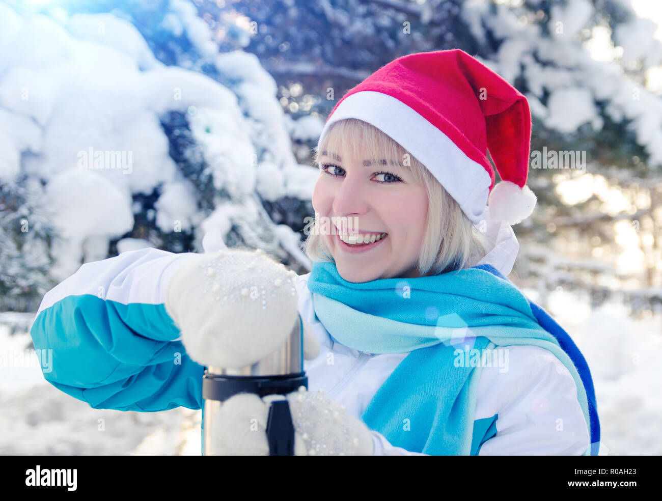 Beautiful Smiling Blonde Girl In A Red Christmas Hat Opens A Thermos With Hot Tea Beautiful Smiling Blonde Girl In A Red Christmas Hat Opens A Thermos With Hot Tea