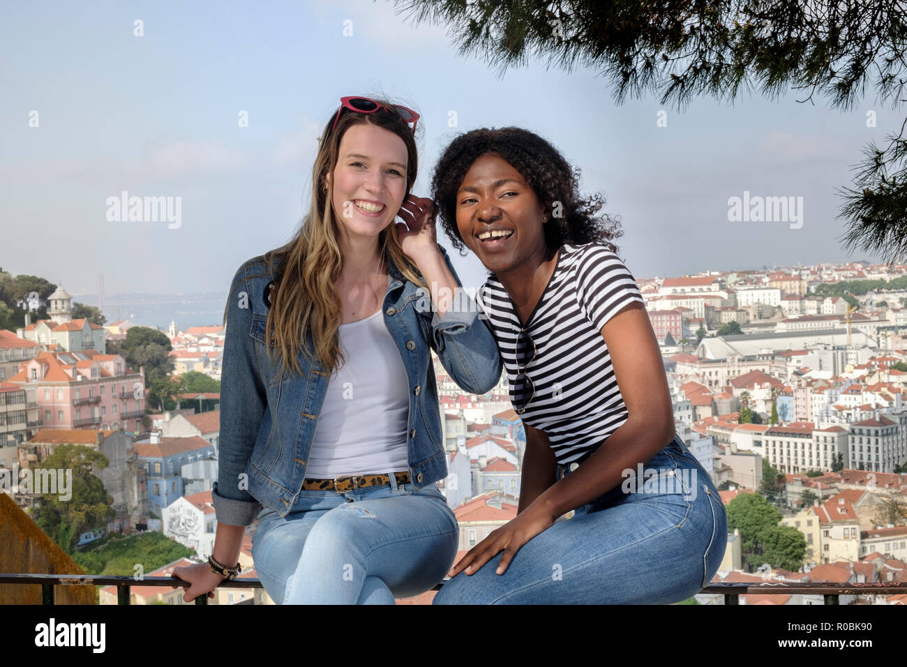 Two young female friends - one African, one European, sitting on a wall on a summer's day with the skyline of Lisbon behind them Stock Photo
