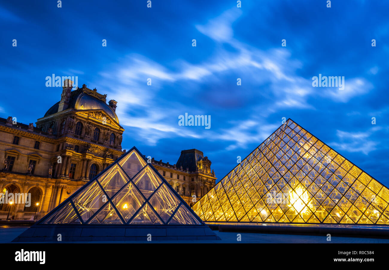 Paris, France - September 30, 2018: Bright glass pyramids inside central square of Louvre museum with long exposure of cloudy twilight blue sky. Stock Photo