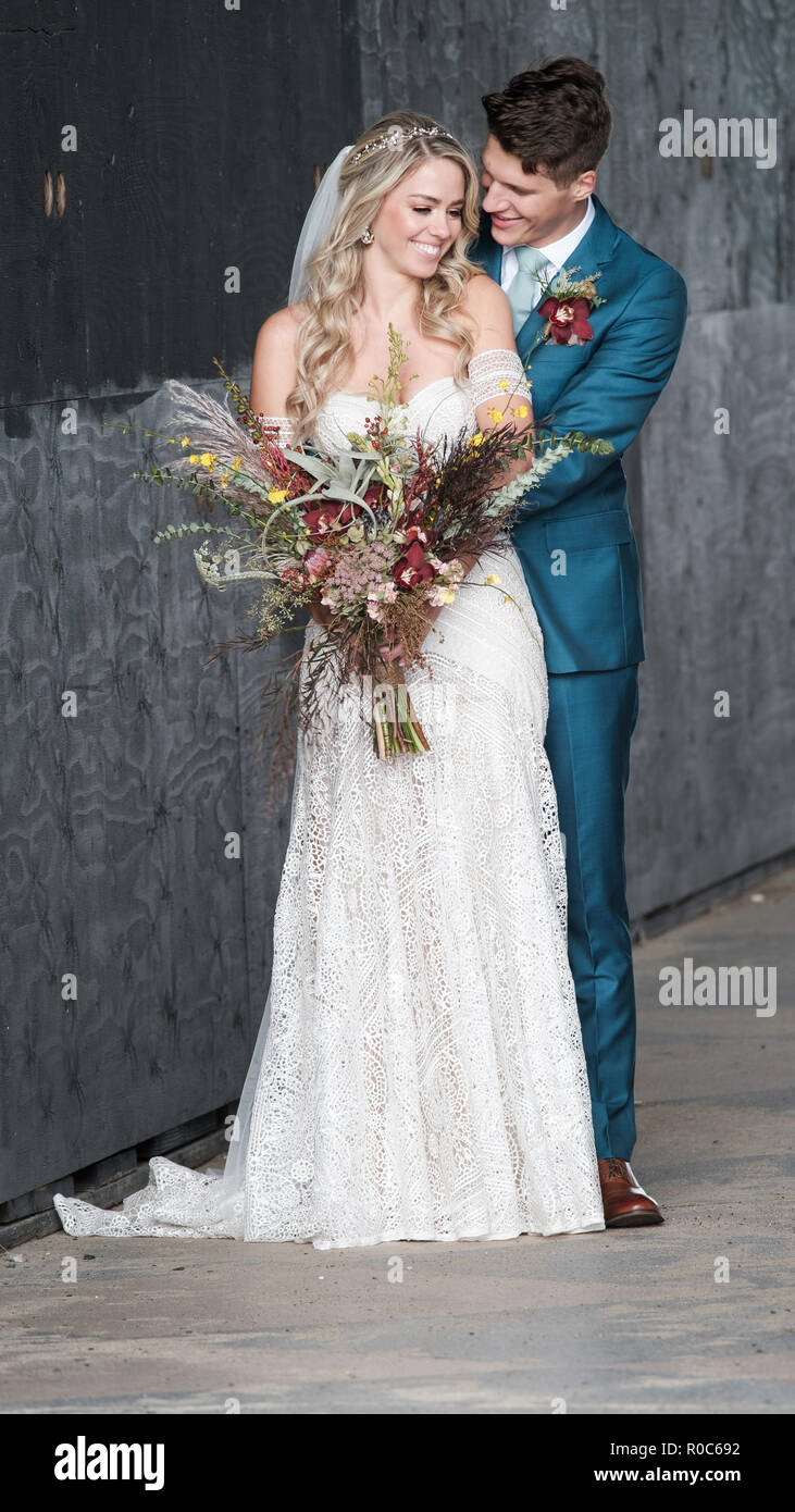 A newly married couple smiling and posing in a loving embrace. The beautiful bride is holding an Autumn bouquet and wearing a white lace wedding dress Stock Photo