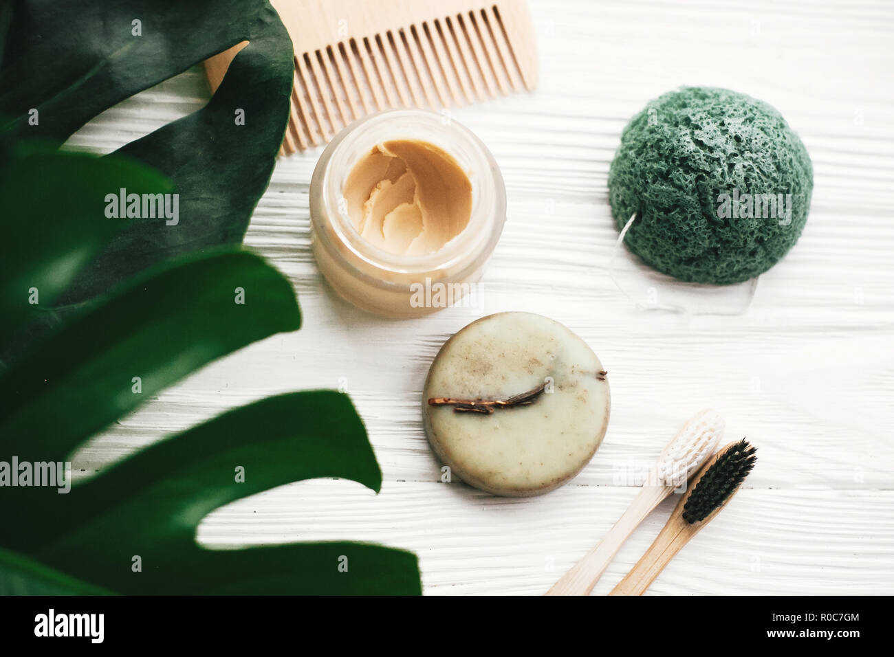 Eco friendly natural bamboo toothbrushes, shampoo bar, toothpaste in glass, wooden brush and konjaku sponge on white wood with green monstera leaves.  Stock Photo