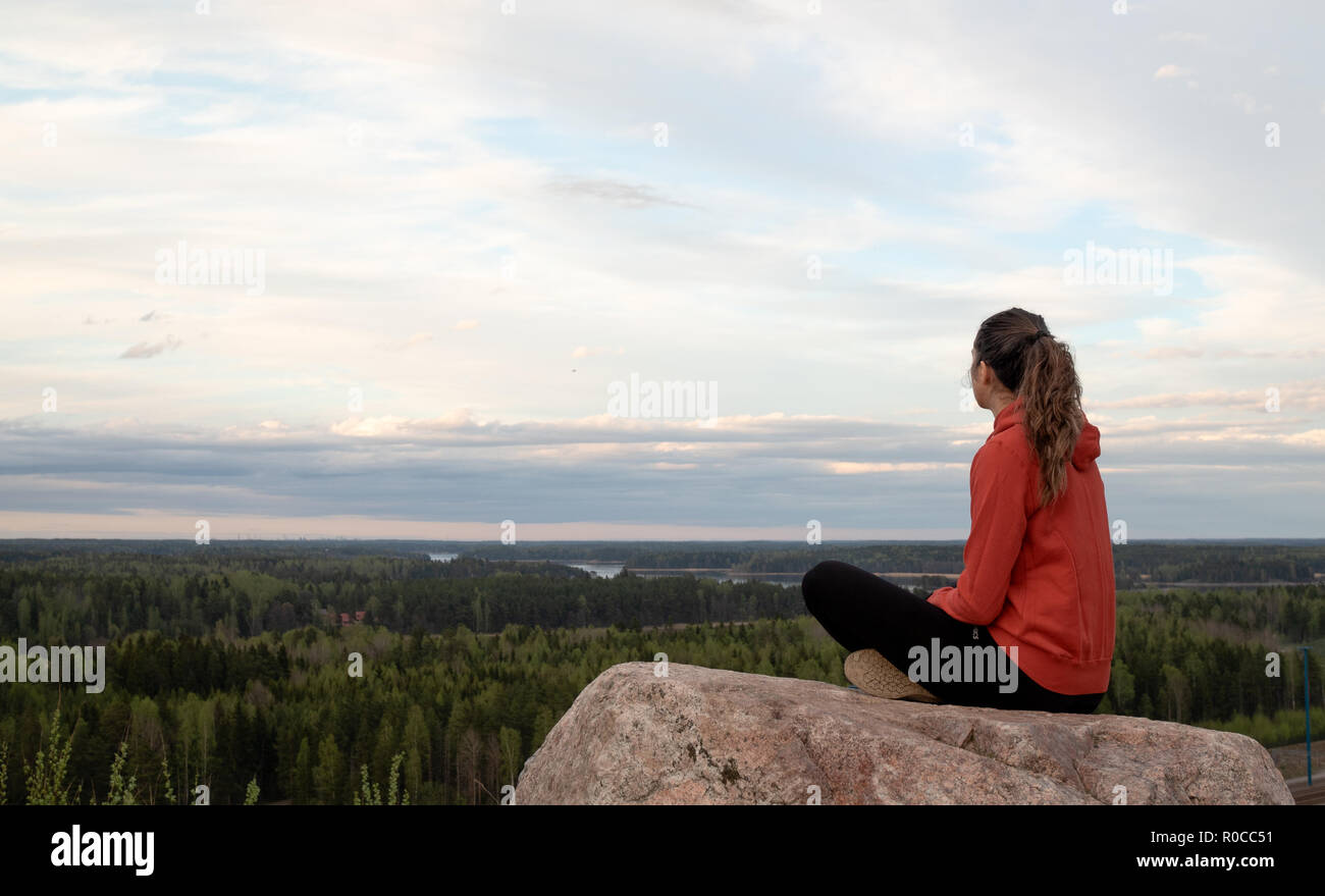 Young beautiful woman doing yoga and mindfulness exercises, while sitting on the edge of a hill and staring to the sunset horizon. Peaceful moment. Stock Photo