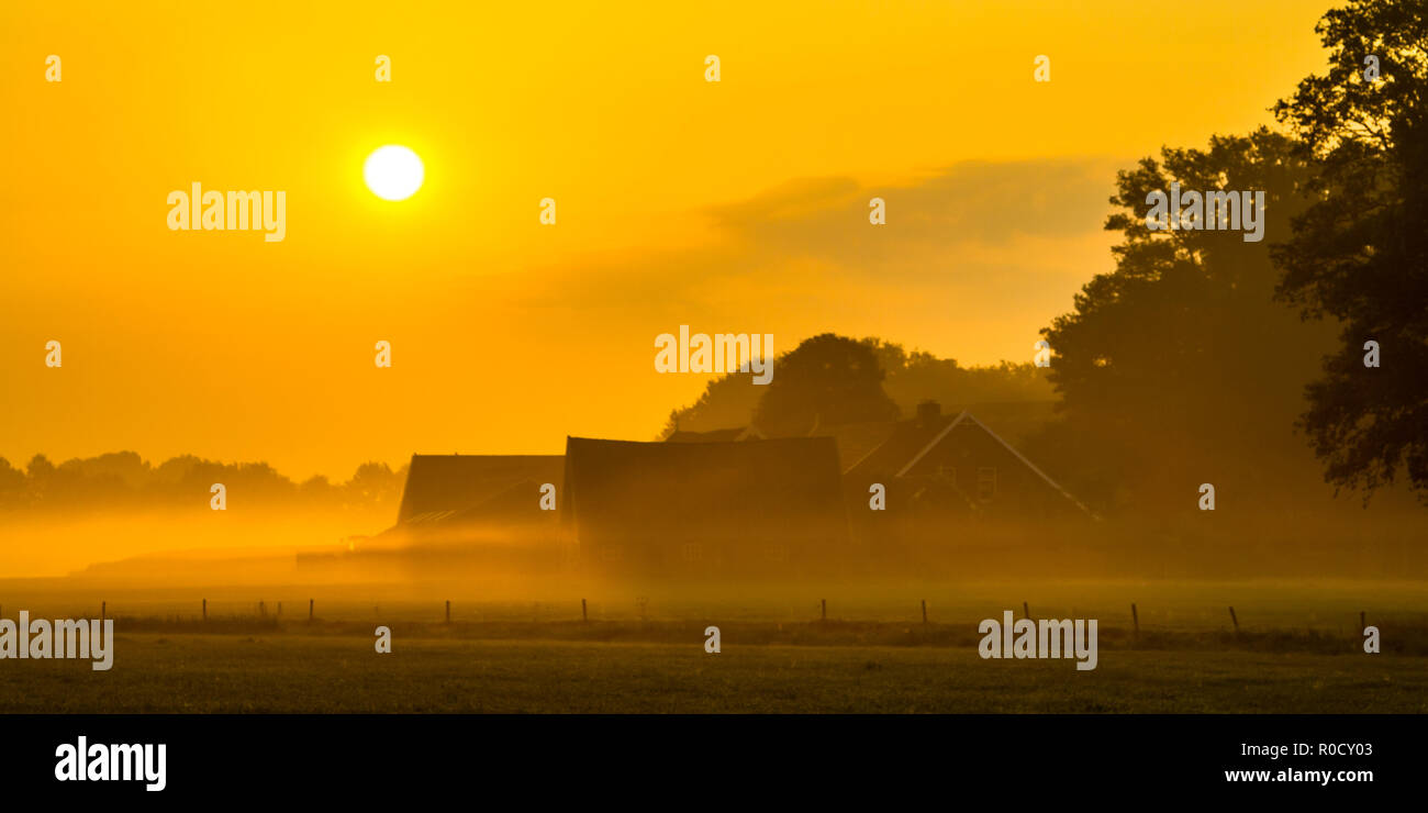 Orange sunrise over misty farmyard with barns and tractor in Twente, Netherlands Stock Photo