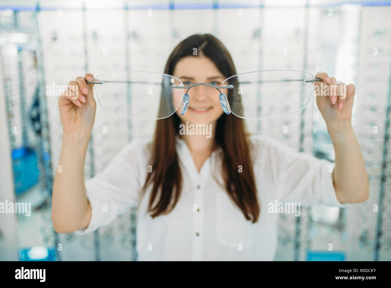 Young woman holds huge decorative glasses, optic store, showcase with spectacles on background. Professional eye care, eyeglasses choice Stock Photo