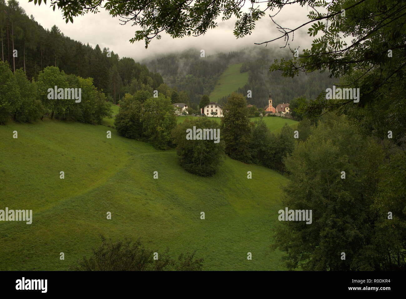 Alpine mountain landscape with typical houses and small church surrounded by lush meadows and dense forest Stock Photo