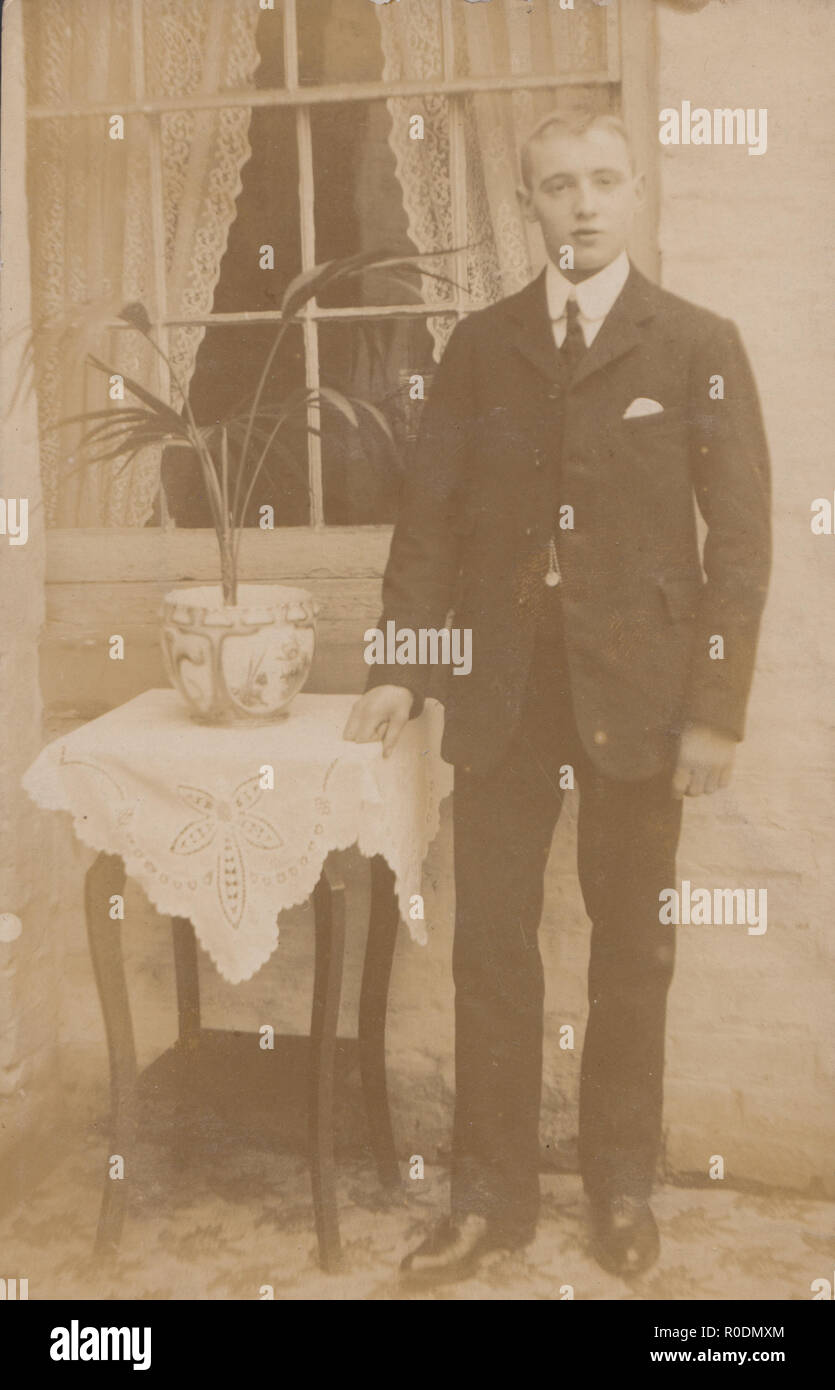 Vintage Photographic Postcard of a Smartly Dressed Young Man Stood Next To a Pot Plant on a Table. Stock Photo